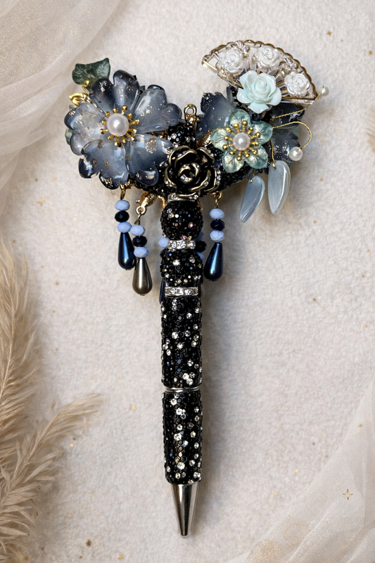 Decorative double-sided black bling pen with large and small flowers, a fan charm with white floral embellishments, and gold accents on a neutral linen background
