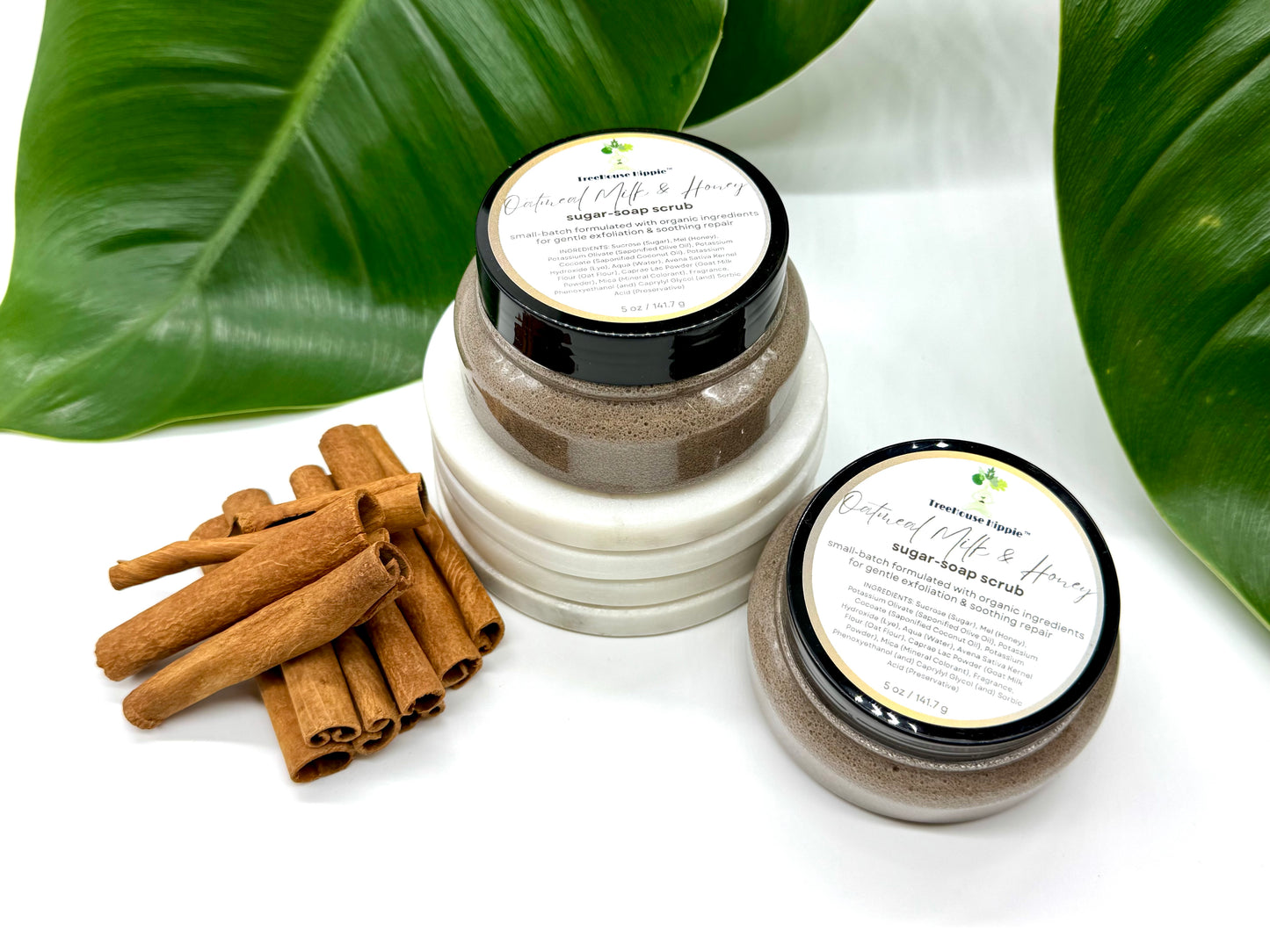 Two of TreeHouse Hippie's 'Oatmeal, Milk, & Honey' scrubs sitting beside cinnamon sticks on a white background with leaf accents.