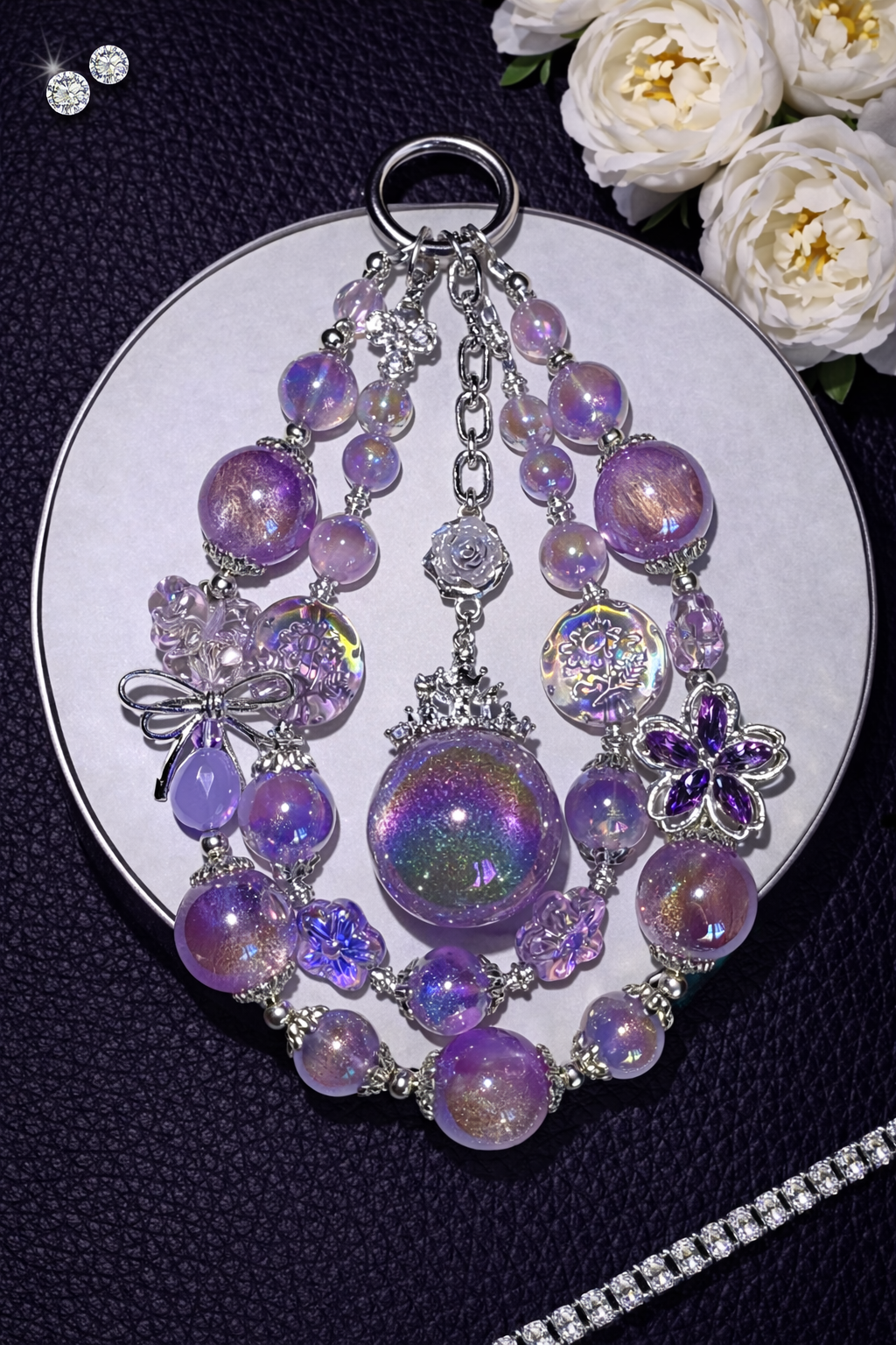 Decorative silver keychain with shimmering chromatic purple beads, a mini purple star, and sparkling accents on a black leather surface with white flowers in the top right corner and a silver chain in the bottom right corner.