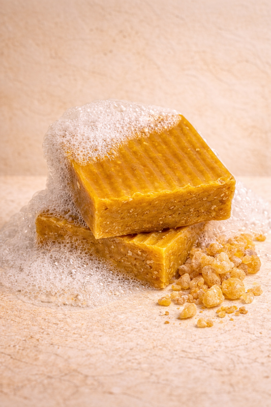 2 bars of TreeHouse Hippie's Sacred Gold organic, handmade soap, stacked one on top of another, covered in bubbles with pieces of amber resin sitting next to them on a neutral beigie background.