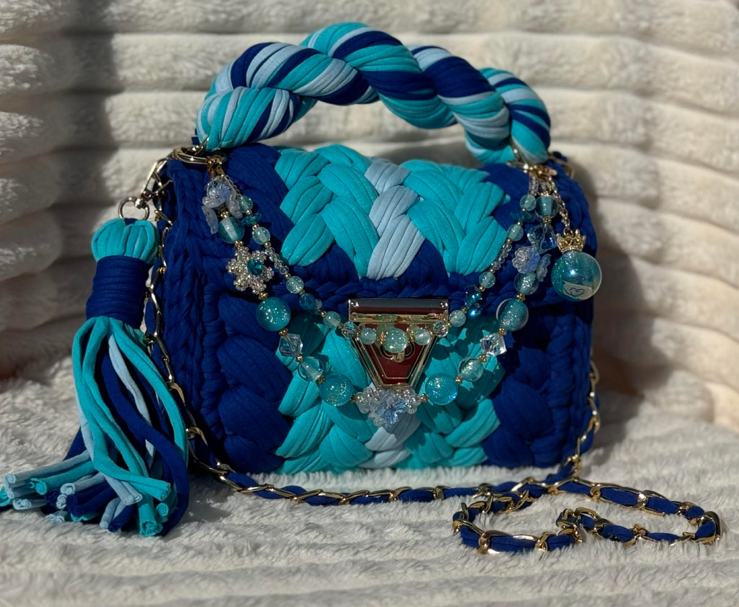 Front view of bag in full sun: Woven Crochet handbag (Royal Blue, Light Blue, and Ice Blue) with tassel and beautiful shimmering keychain sitting on a white, textured surface.