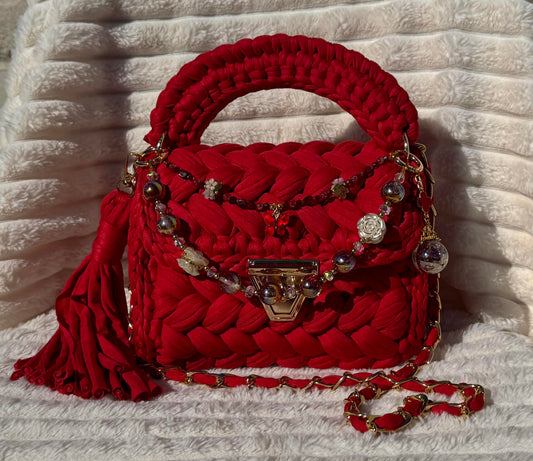 Front view in full sun: Red woven Crochet handbag with decorative keychain chain and tassel on a textured White background. 