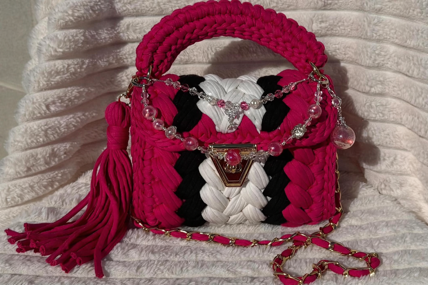 Front View of the bag in full sunlight showing: Hot Pink, Black, and White Crochet woven handbag with Keychain and decorative elements sitting on a textured White surface
