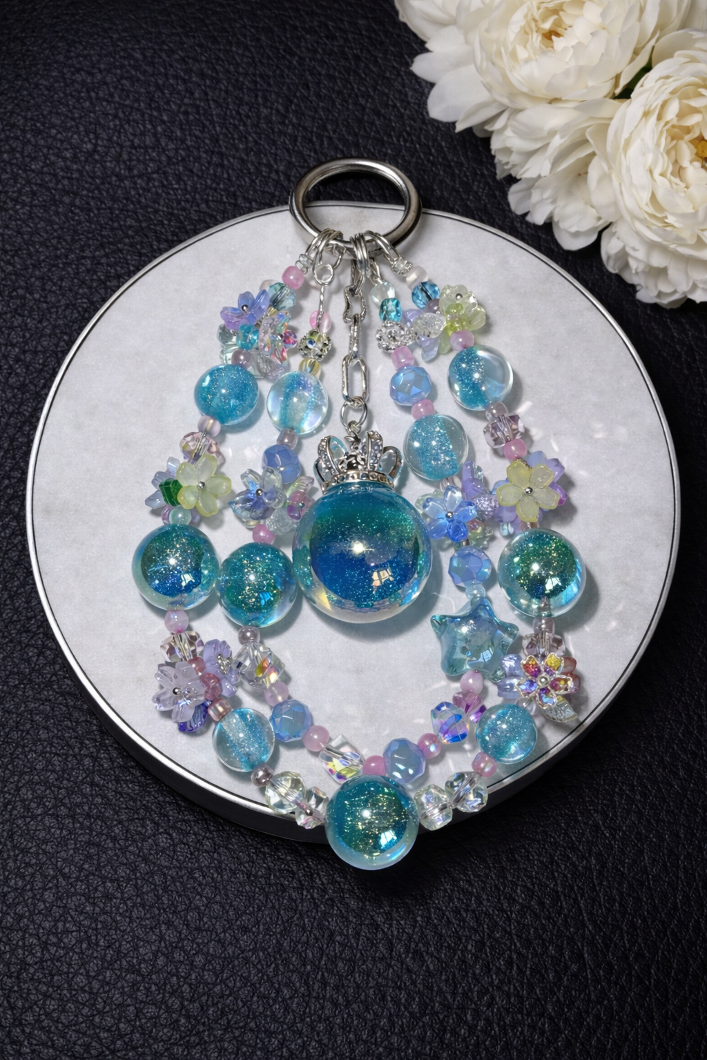 Decorative silver keychain with shimmering blue-green beads, star charms, and multicolor floral accents on a dark leather surface with white flowers in the top right corner. 
