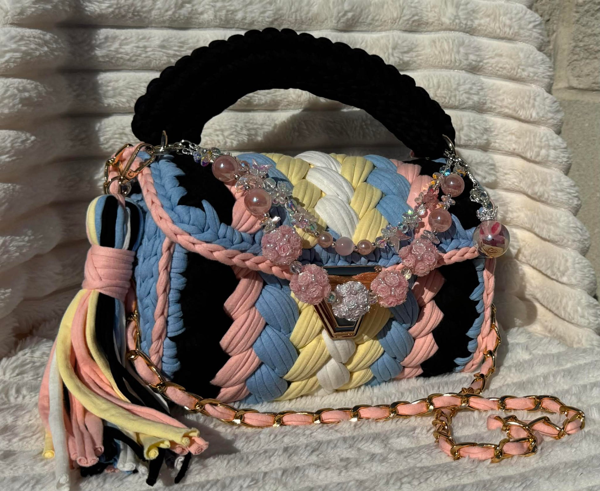 Side/Front view in full sun: Multicolored woven Crochet handbag with decorative keychain and tassel sitting on a textured white background. Bag colors include: Black, Light Blue, Pink, Soft Yellow, and White. 