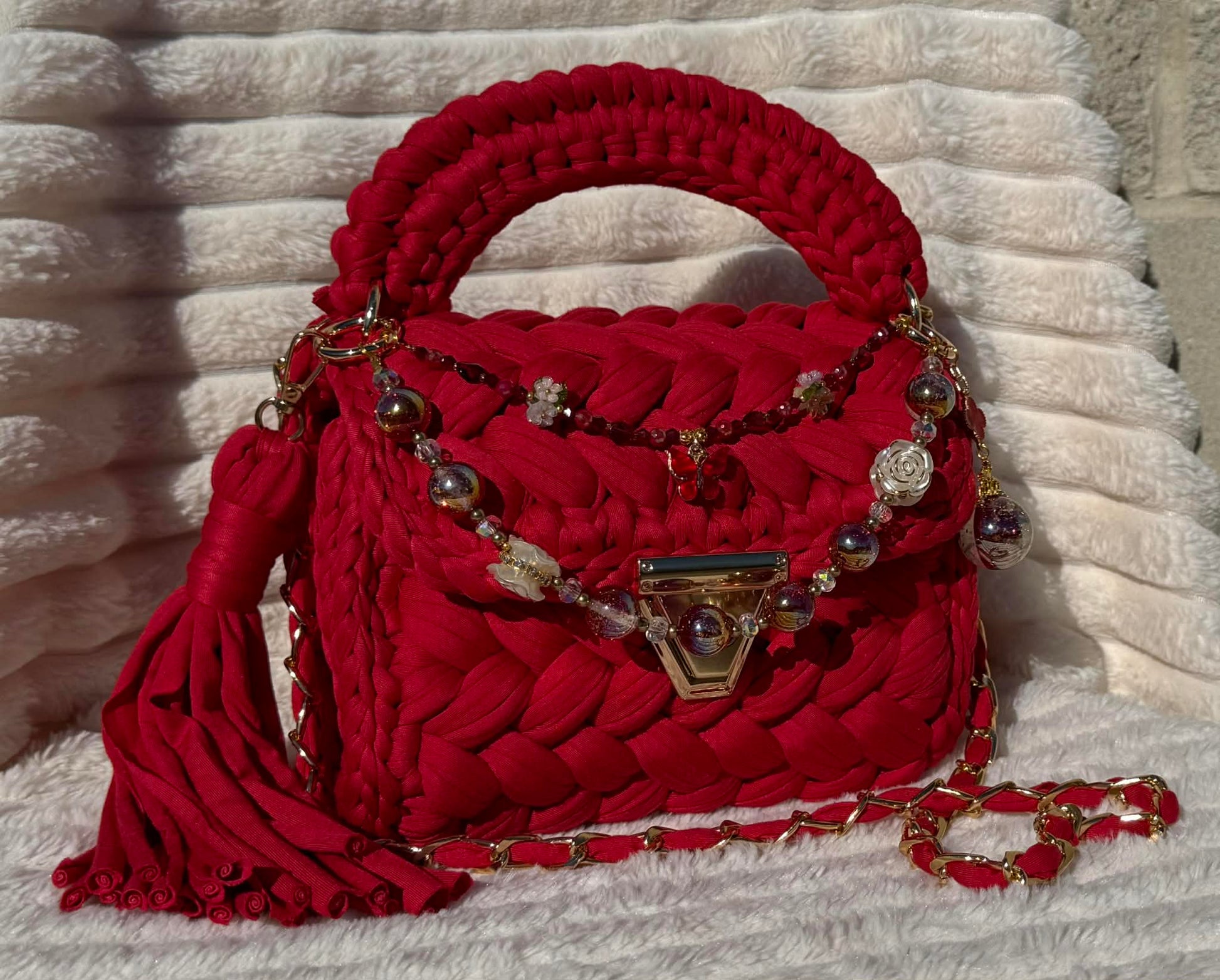 Side/Front view in full sun: Red woven Crochet handbag with decorative keychain chain and tassel on a textured White background. 