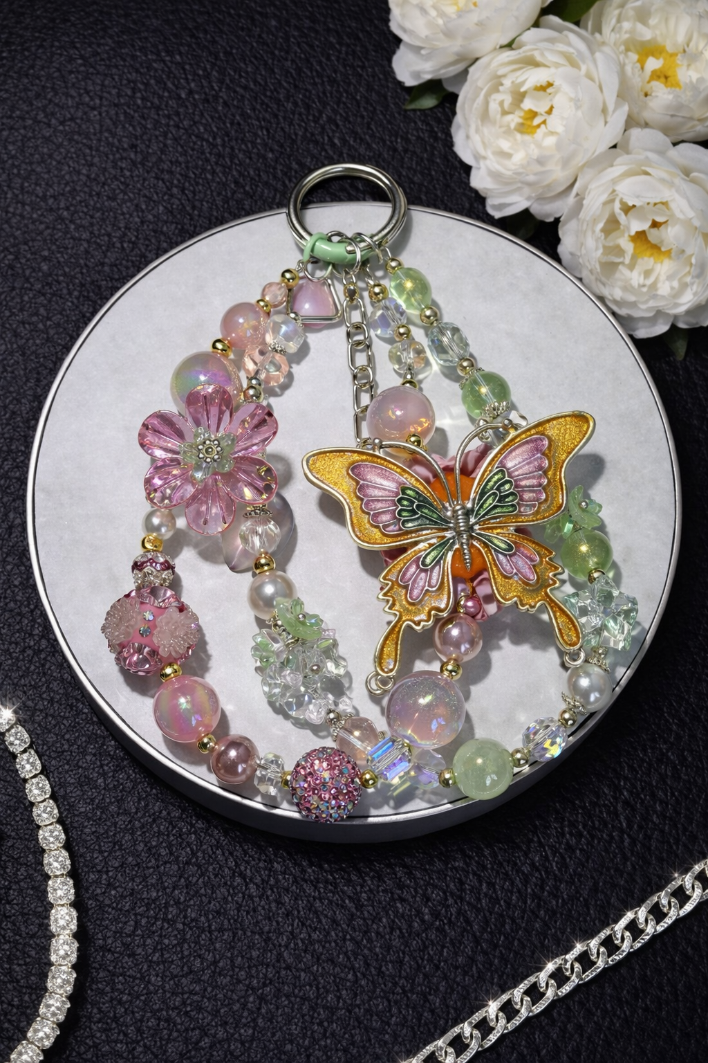 Decorative silver keychain with pink and green beads and a large butterfly charm on a round white tray, on a black leather surface with flowers in the top right corner. 