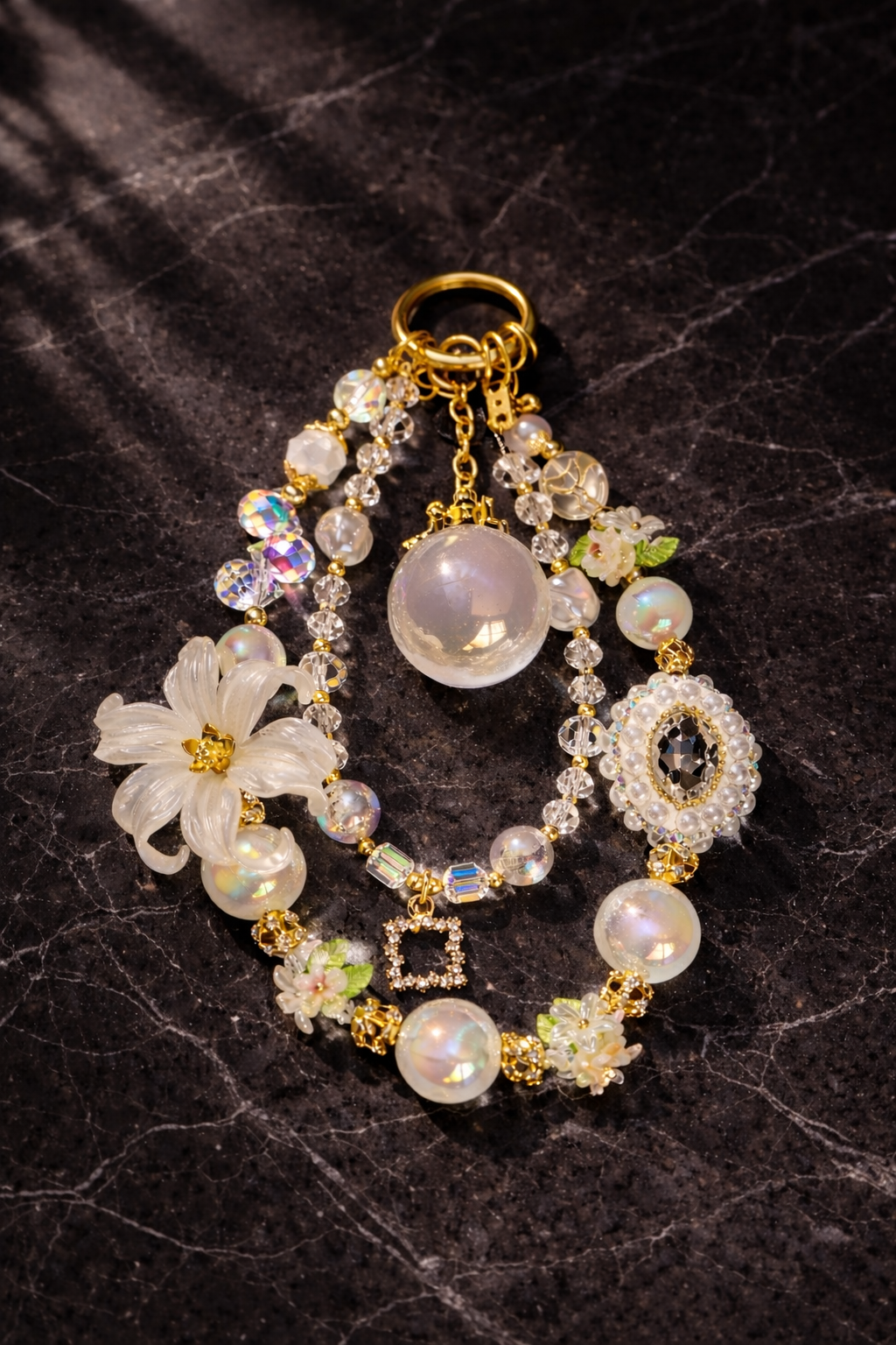 Decorative gold and white keychain with pearl beads, a large flower charm, and shimmering crystals on a dark textured marble background. 