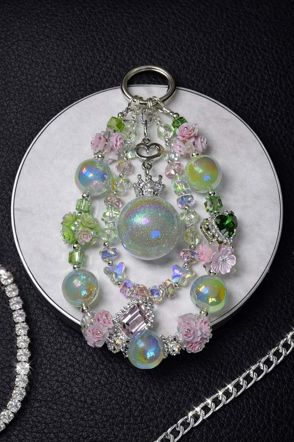 Decorative silver keychain with  green and yellow shimmering beads and pink flower accents on a white tray, sitting on a black leather background. 