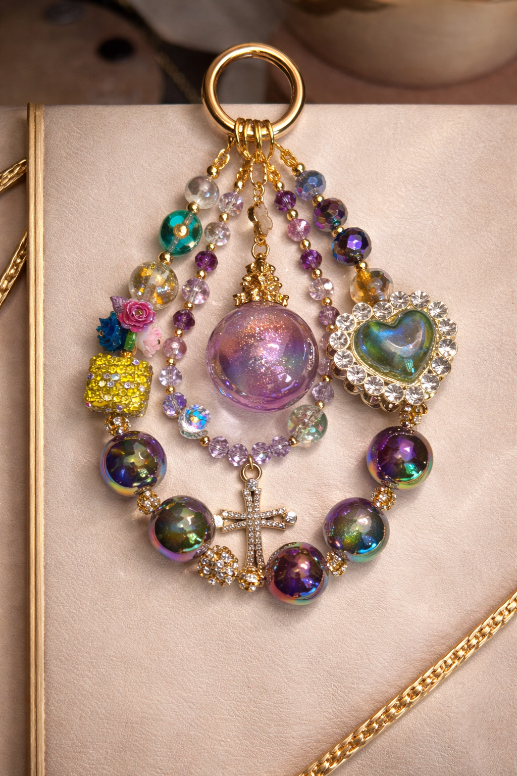 Colorful decorative keychain with deep purple shimmering beads, a large light green heart charm, and a cross in the  middle on a soft, beige surface with gold trim.