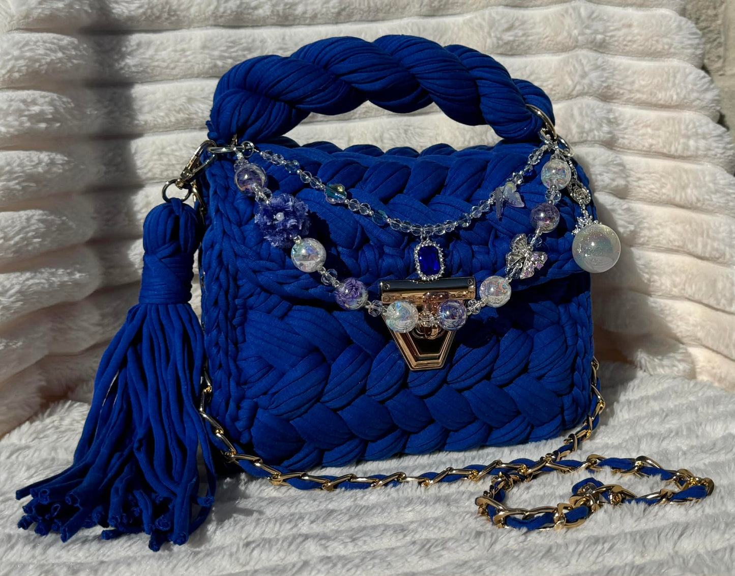 Front View of the bag in full sun: Royal/Indigo Blue Crochet woven handbag with decorative keychain chain and tassel on a textured White background surface.