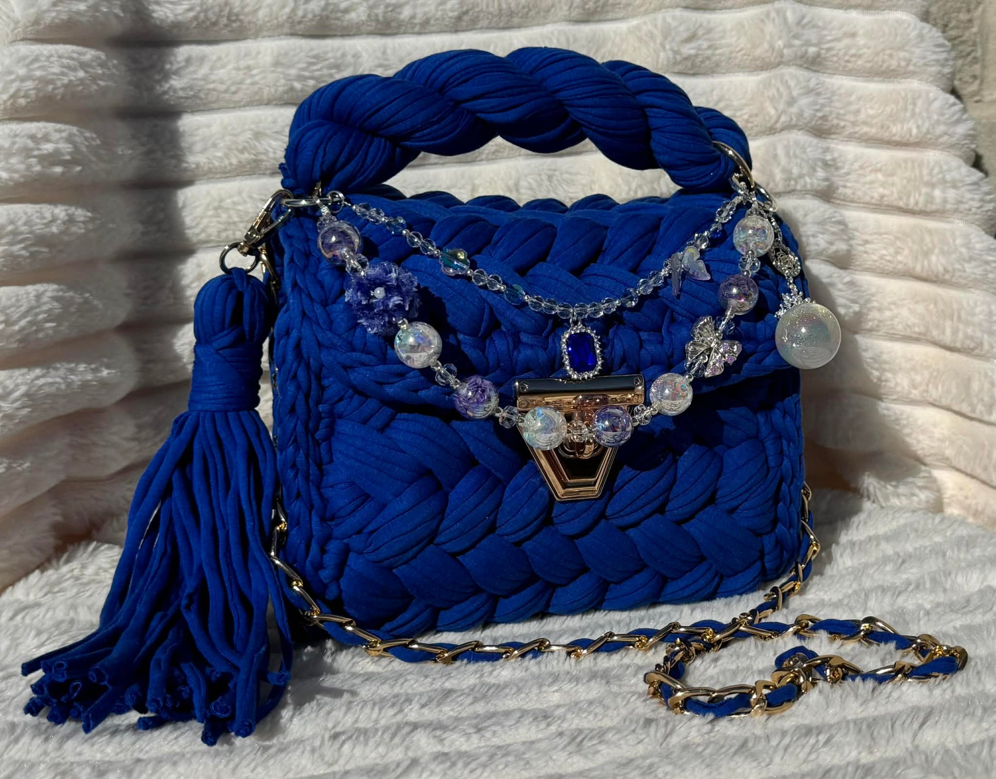 Front View of the bag in full sun: Royal/Indigo Blue Crochet woven handbag with decorative keychain chain and tassel on a textured White background surface.
