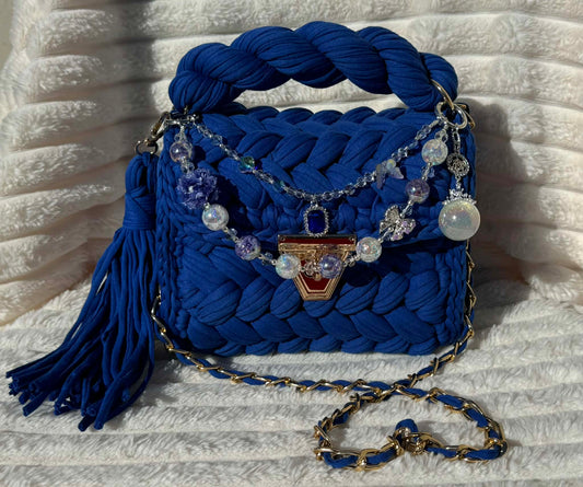 Front View of the bag in full sun: Royal/Indigo Blue Crochet woven handbag with decorative keychain chain and tassel on a textured White background surface.