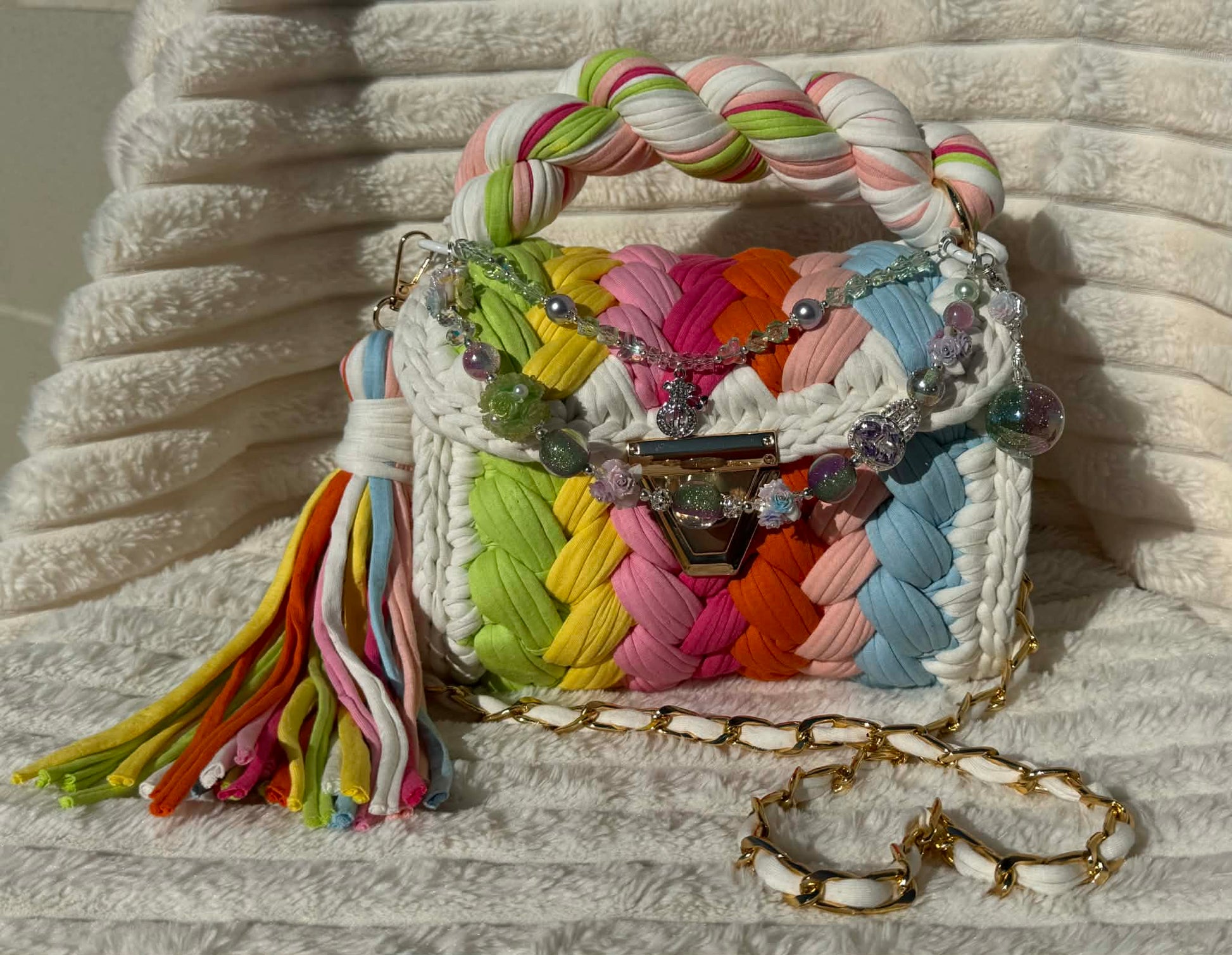 Front view of the bag in full sun: Colorful woven Crochet handbag with pastel rainbow pattern, tassel, and shimmering keychain sitting on a white, textured surface. Pastel Rainbow pattern includes: Lime Green, Orange, Yellow, Light Blue, Pink, and White. 