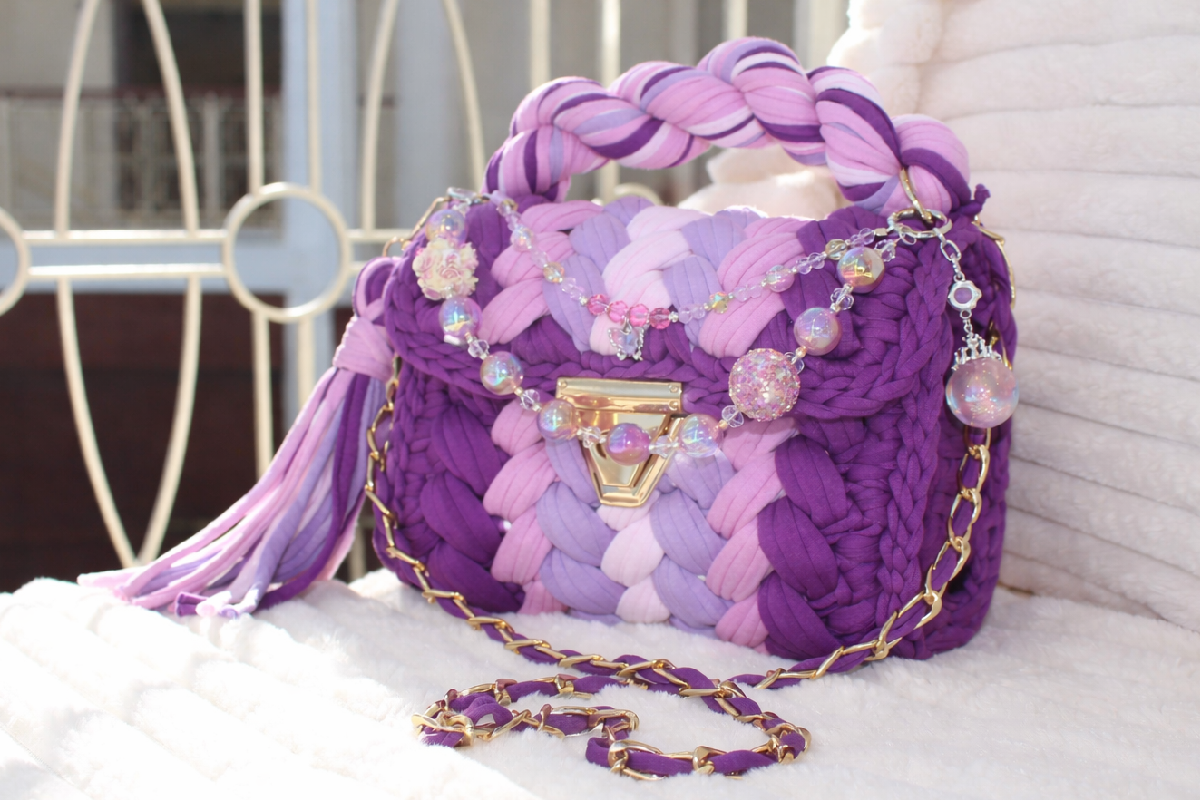 Side/Front view of bag in natural lighting: Colorful woven Crochet handbag (Purple, Lavender, Lilac, Periwinkle, & Blush) with tassel and beautiful shimmering keychain sitting on a white, textured surface. 