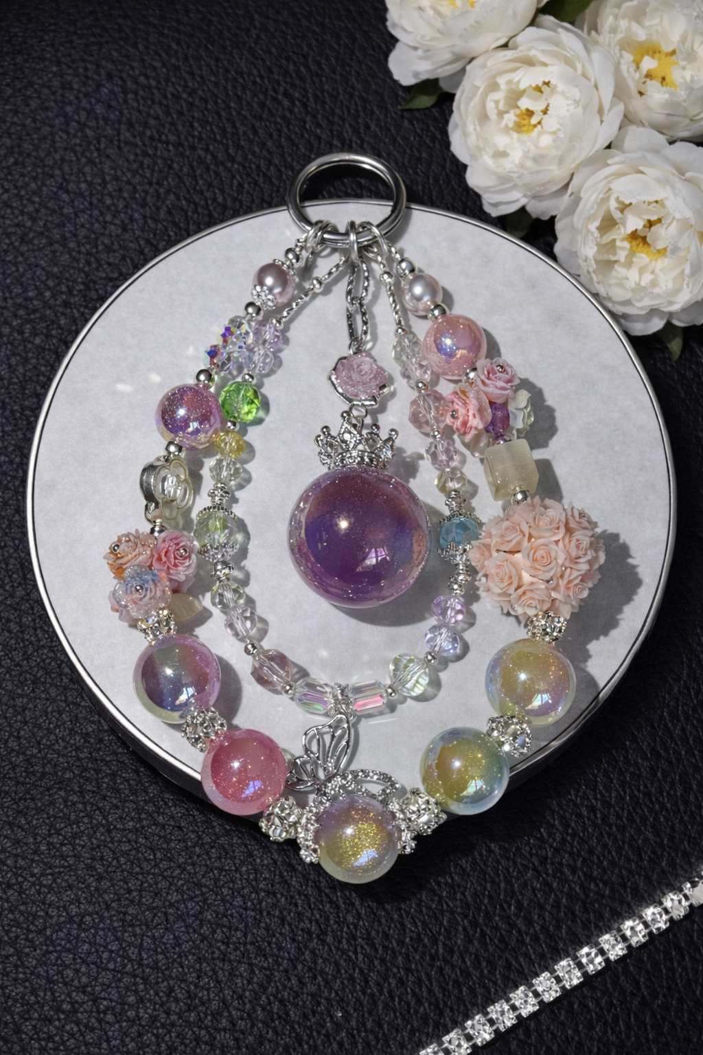 Decorative silver keychain with shimmering pink, yellow, and purple beads, multicolor floral accents, and a butterfly pendant on a dark leather surface with white flowers in the top right corner and a silver chain in the bottom right corner. 