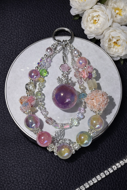 Decorative silver keychain with shimmering pink, yellow, and purple beads, multicolor floral accents, and a butterfly pendant on a dark leather surface with white flowers in the top right corner and a silver chain in the bottom right corner. 