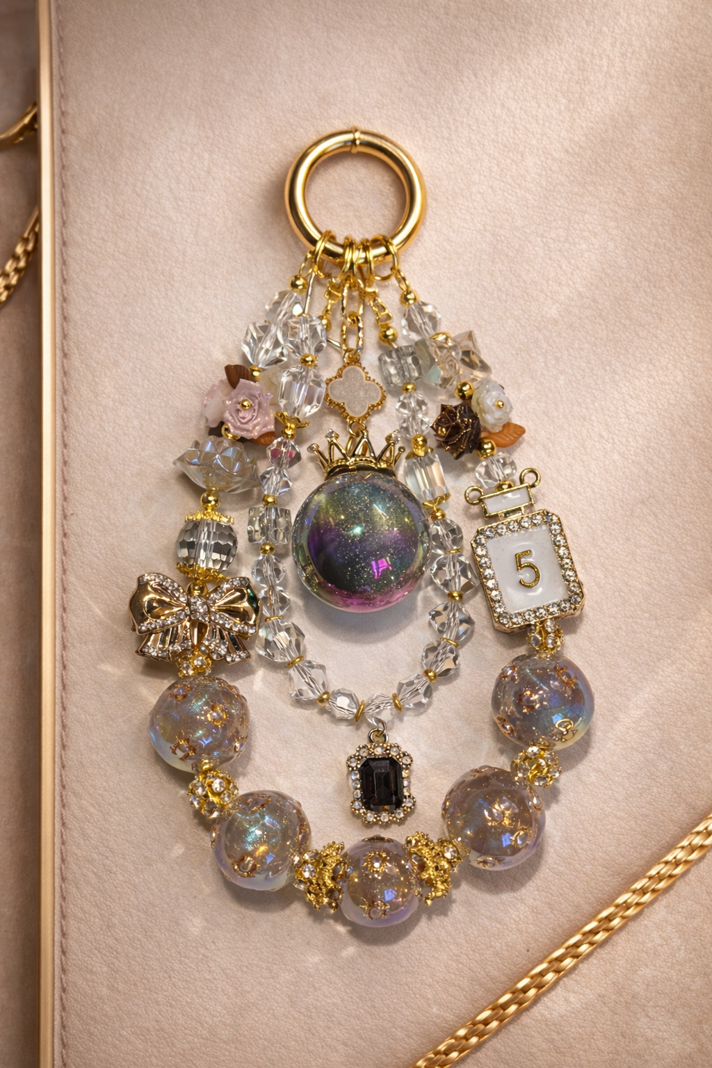 Decorative keychain with iridescent glass beads, floral accents, and a perfume bottle charm on a soft, beige background with gold trim. 
