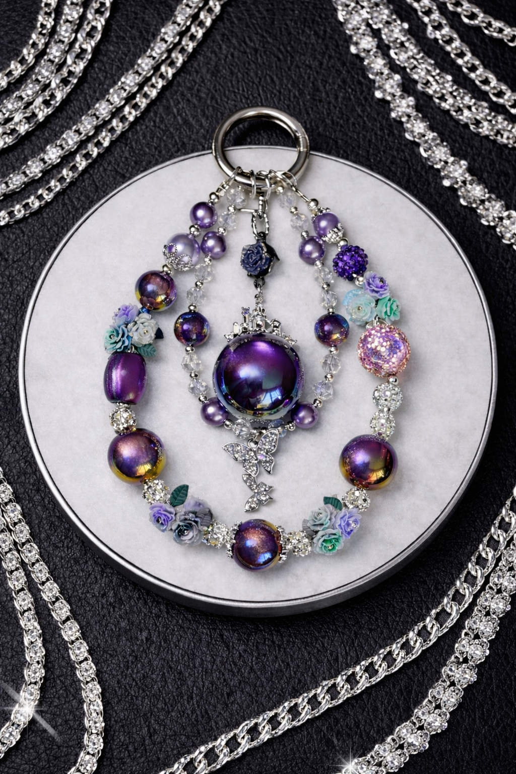 Decorative silver keychain with dark purple beads, flower accents, and a butterfly pendant on a white tray, sitting on a black leather background with silver chains in the corners. 