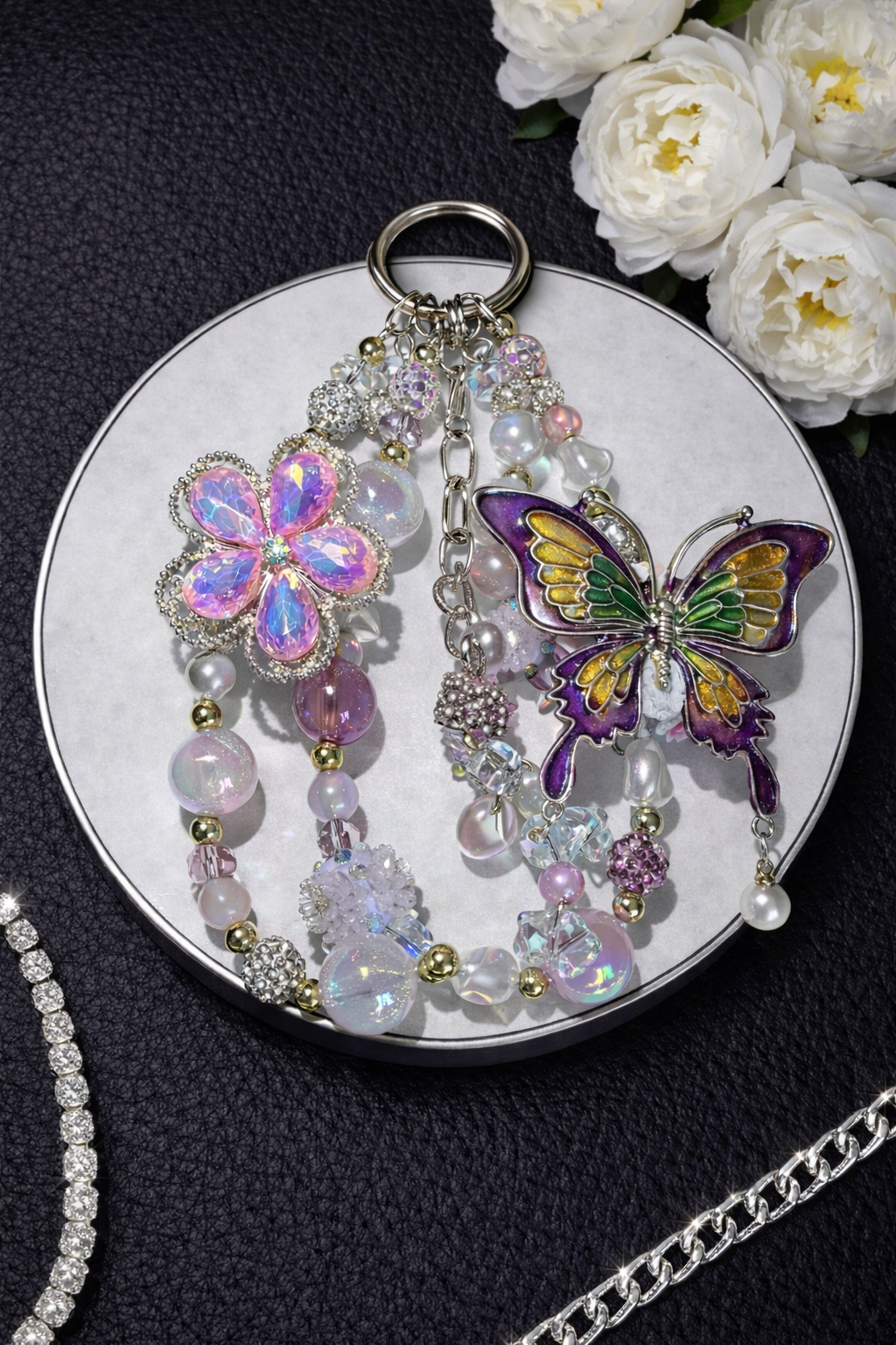 Decorative silver keychain with shimmering white glass beads, a large butterfly charm, and a large flower charm on a black leather surface with flowers in the top right corner. 