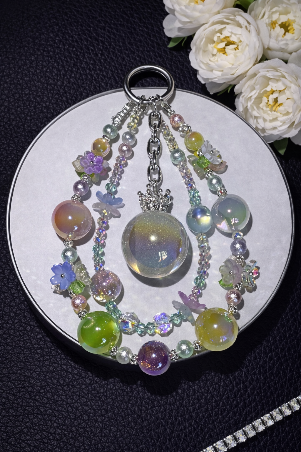 Decorative silver keychain with green, purple, yellow, pink, & blue glass beads and multicolor floral accents on a dark leather surface with white flowers in the top right corner and a silver chain in the bottom right corner. 