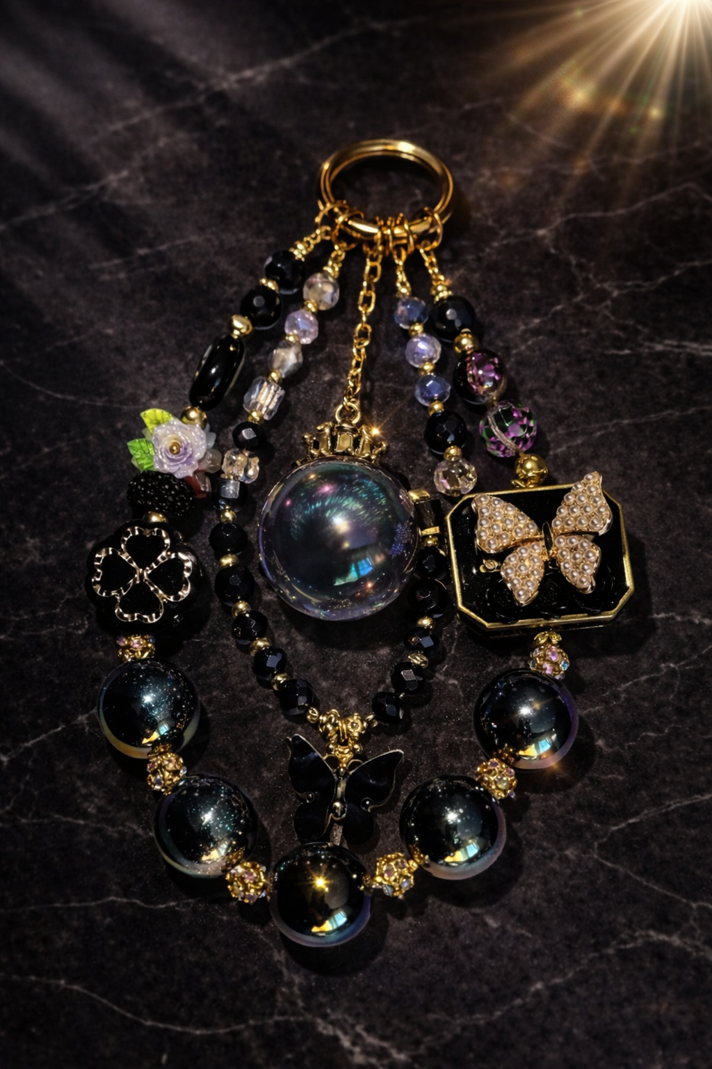 Decorative black and gold keychain with pearl black beads, a black clover charm, and a black butterfly pendant  on a dark marble background. 