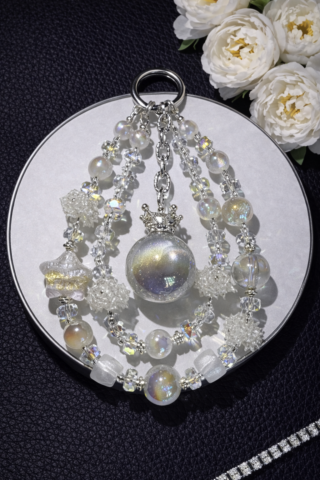 Decorative silver keychain with shimmering white-gold pearl beads, white pearl flower accents, and a icey white star bead on a black leather surface with white flowers in the top right corner and a silver chain in the bottom right corner. 