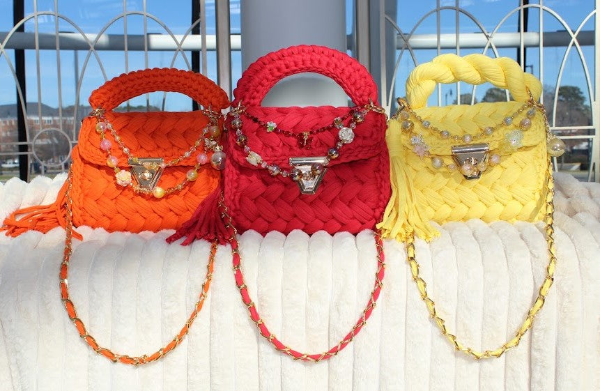 Three colorful handbags (Orange, Red, and Yellow) with decorative keychains and accessories on a textured white surface.
