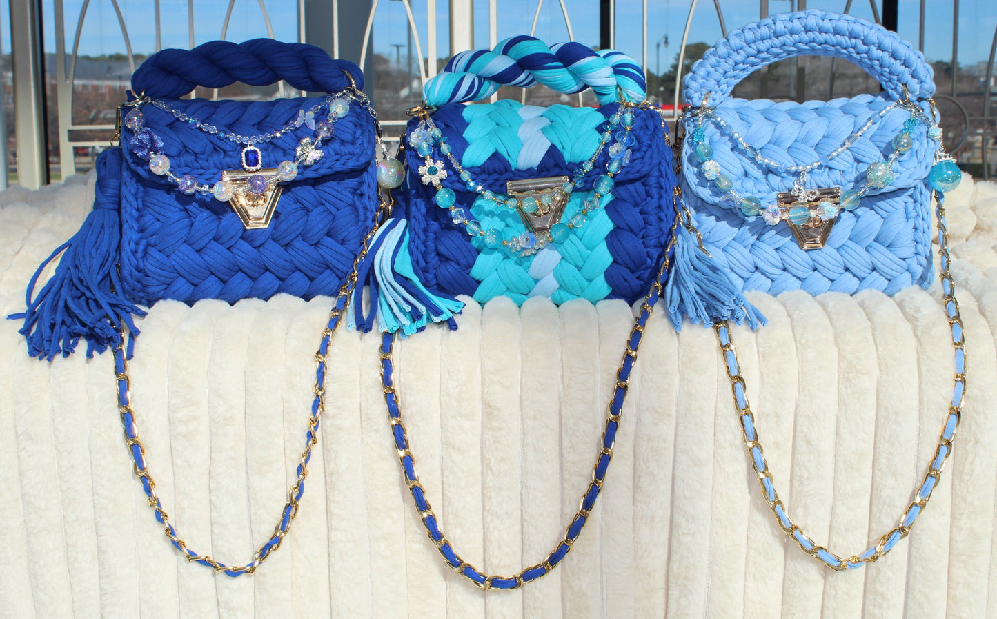 Three Blue handbags in different shades of Blue with decorative keychains sitting on a White textured surface.