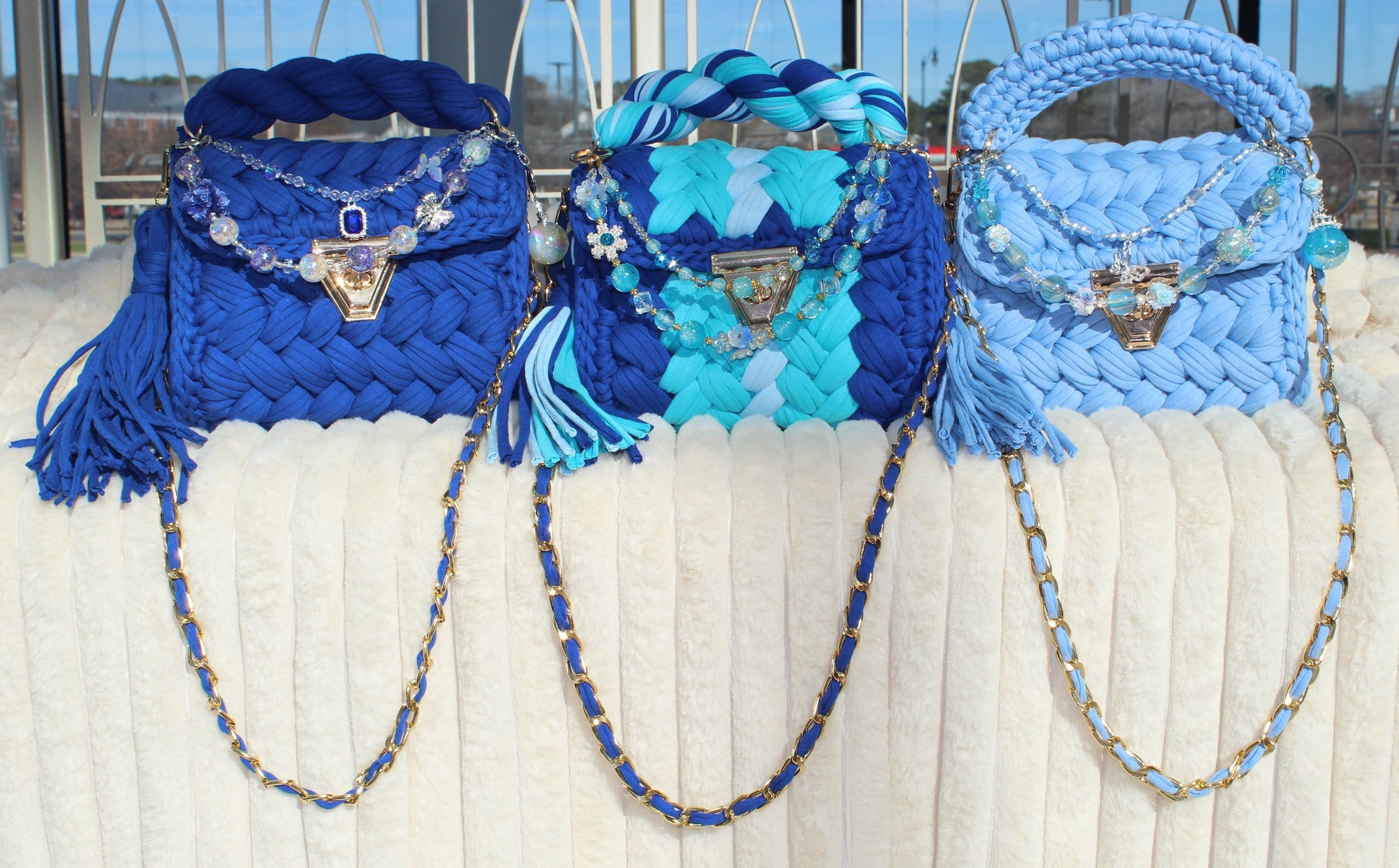 Three Blue handbags in different shades of Blue with decorative keychains sitting on a White textured surface.