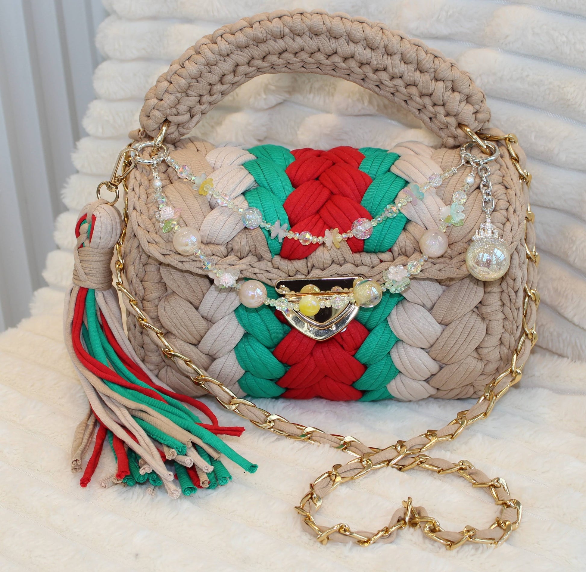 Front view of bag in natural lighting: Shades of Green, Red, Tan, and Nude with decorative blingy keychain and tassel sitting on a white tectured surface and background. 