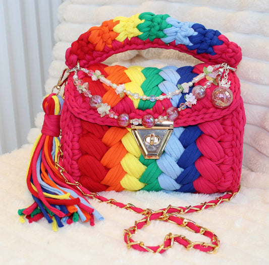 Front of the bag in natural lighting: Colorful Crochet woven handbag with rainbow pattern and beautiful decorative keychain on a white surface. Rainbow pattern includes: Hot Pink, Red, Orange, Yellow, Green, Light Blue, and Dark Blue. 