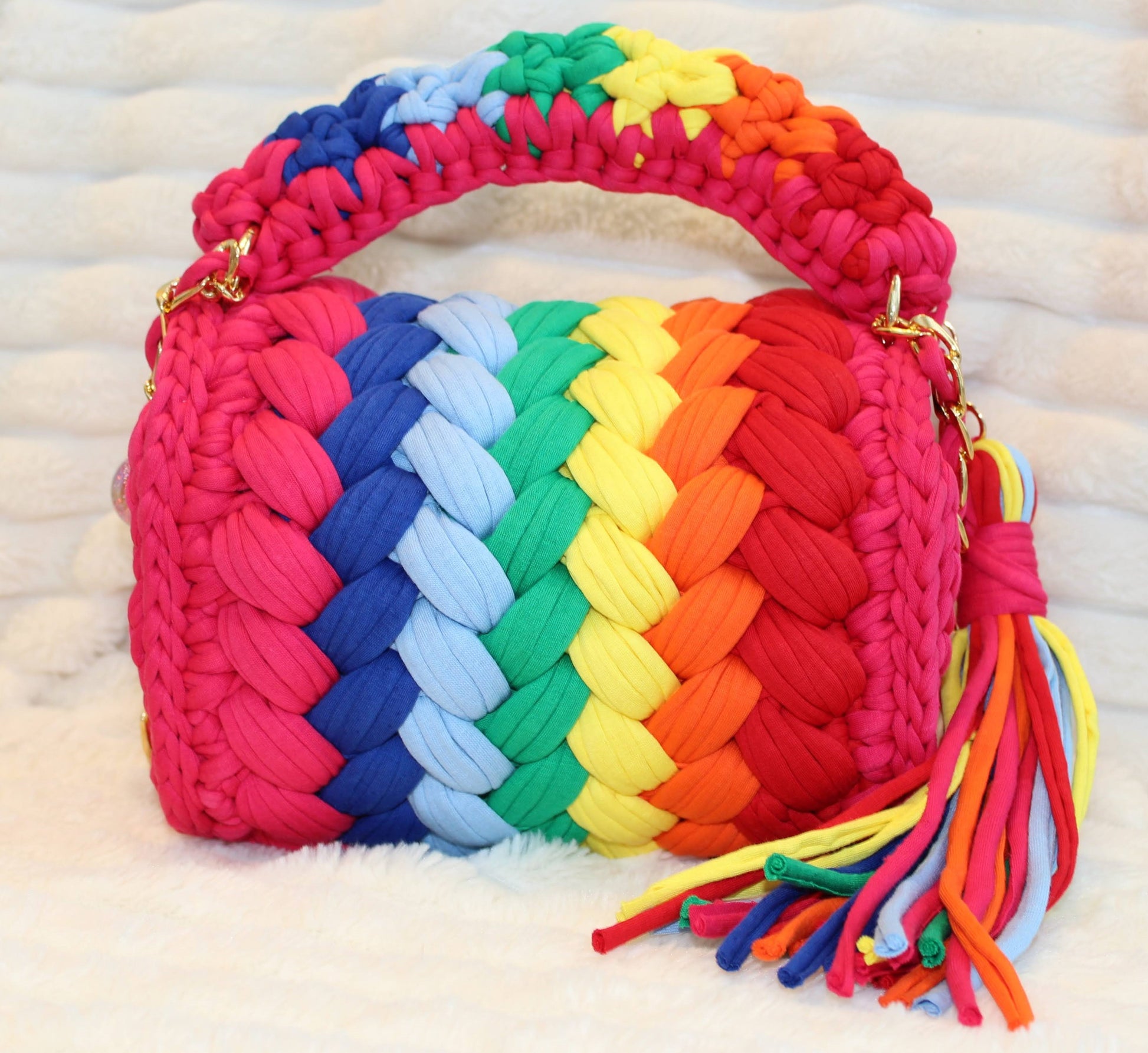 Back of the bag in natural lighting: Colorful Crochet woven handbag with tassel in rainbow pattern on a white surface. Rainbow pattern includes: Hot Pink, Red, Orange, Yellow, Green, Light Blue, and Dark Blue. 