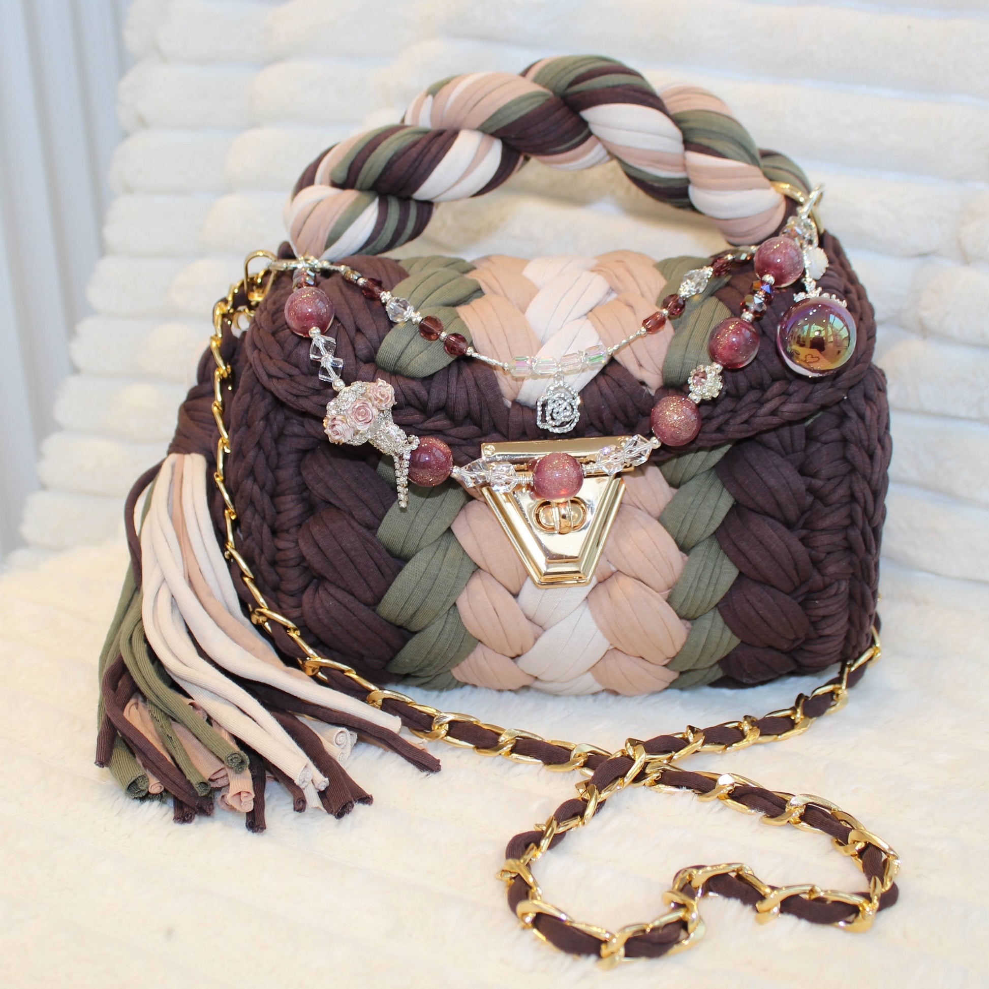 Front view of bag in natural lighting: Colorful woven Crochet handbag (Brown, Tan, Moss Green, and Beige) with tassel and beautiful shimmering keychain sitting on a white, textured surface. 
