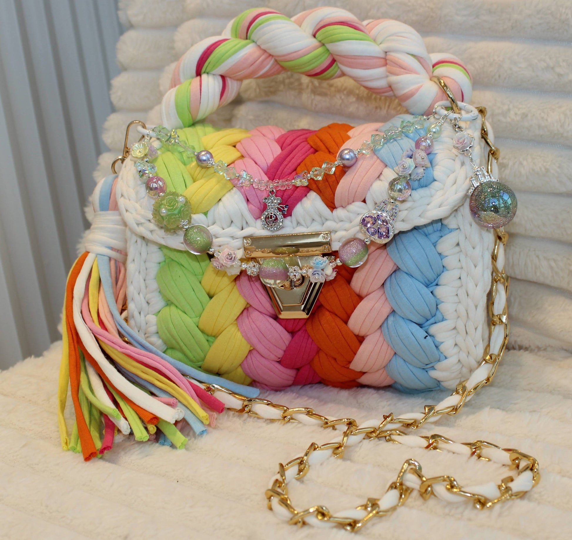 Front view of bag in natural lighting: Colorful woven Crochet handbag with pastel rainbow pattern, tassel, and shimmering keychain sitting on a white, textured surface. Pastel Rainbow pattern includes: Lime Green, Orange, Yellow, Light Blue, Pink, and White. 