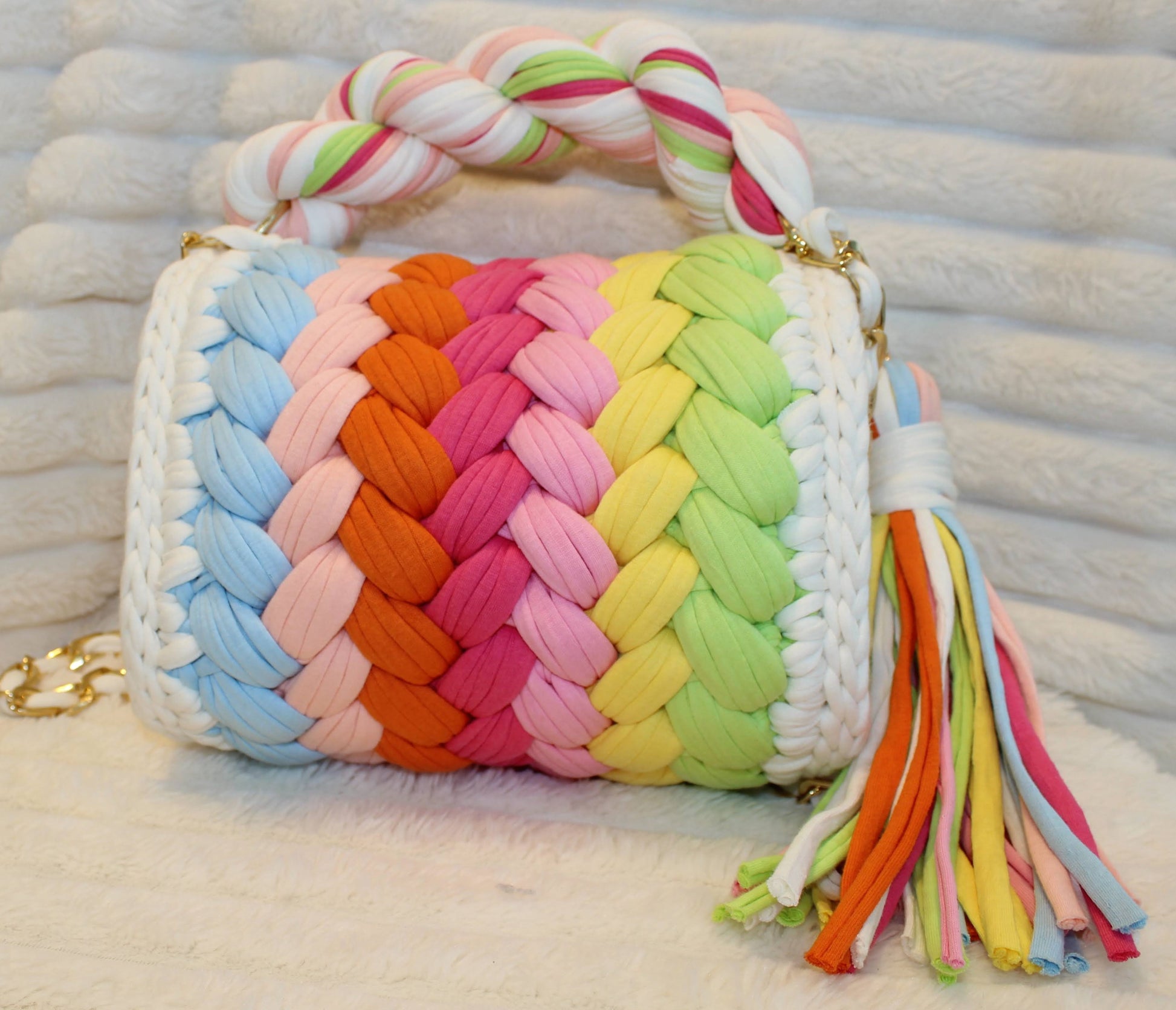 Back view of bag in natural lighting: Colorful woven Crochet handbag with pastel rainbow pattern and tassel sitting on a textured white surface. Pastel Rainbow pattern includes: Lime Green, Orange, Yellow, Light Blue, Pink, and White. 