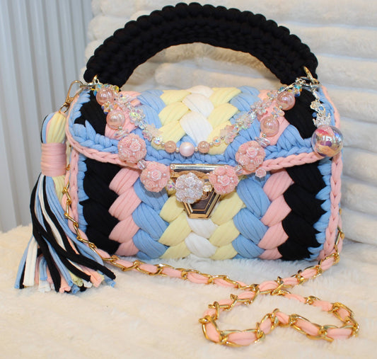 Front view in natural lighting: Multicolored woven Crochet handbag with decorative keychain and tassel sitting on a textured white background. Bag colors include: Black, Light Blue, Pink, Soft Yellow, and White. 