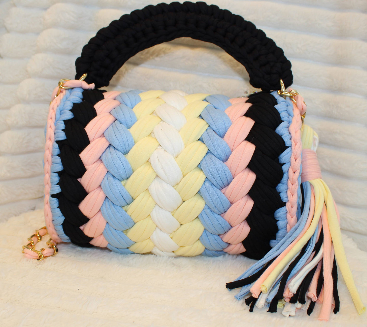 Back view in natural lighting: Multicolored woven Crochet handbag with tassel sitting on a textured white background. Bag colors include: Black, Light Blue, Pink, Soft Yellow, and White. 