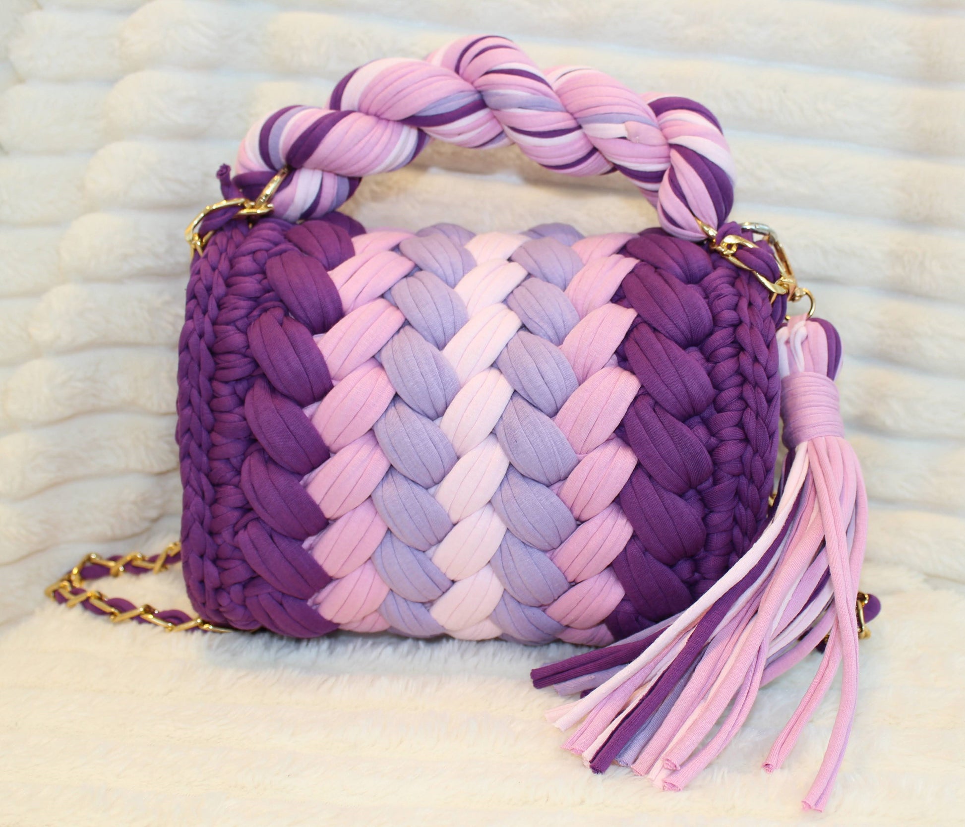 Back view of bag in natural lighting: Colorful woven Crochet handbag (Purple, Lavender, Lilac, Periwinkle, & Blush) with tassel, sitting on White textured surface. 