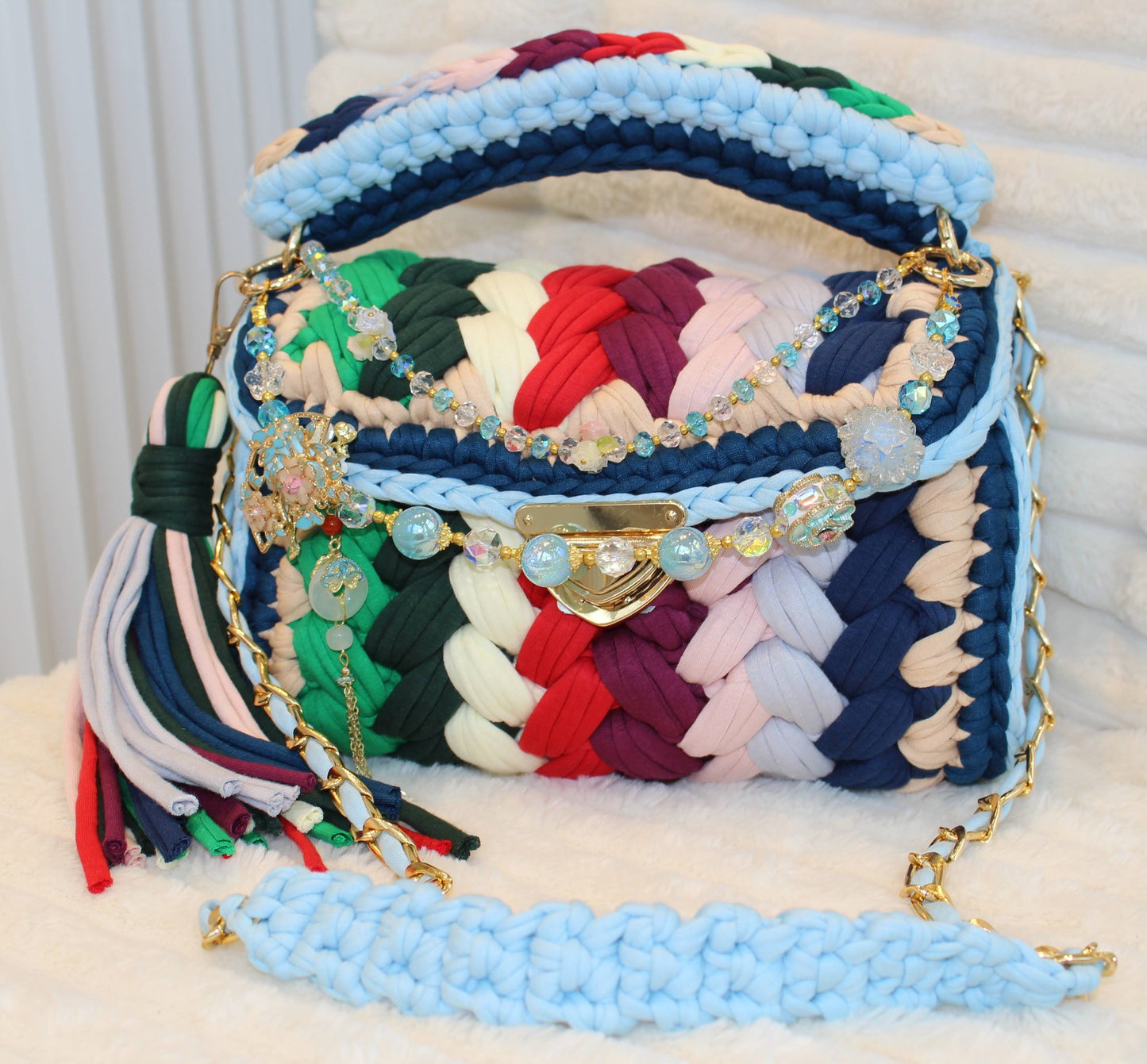 Front view of bag: Colorful handbag in Crochet woven design with keychain, tassel, and decorative elements on a White textured surface. Bag colors include: Tan, Blue, Green, Forest Green, Red, Burgundy, Soft Yellow, Nude, Light Blue, and Dark Blue. 