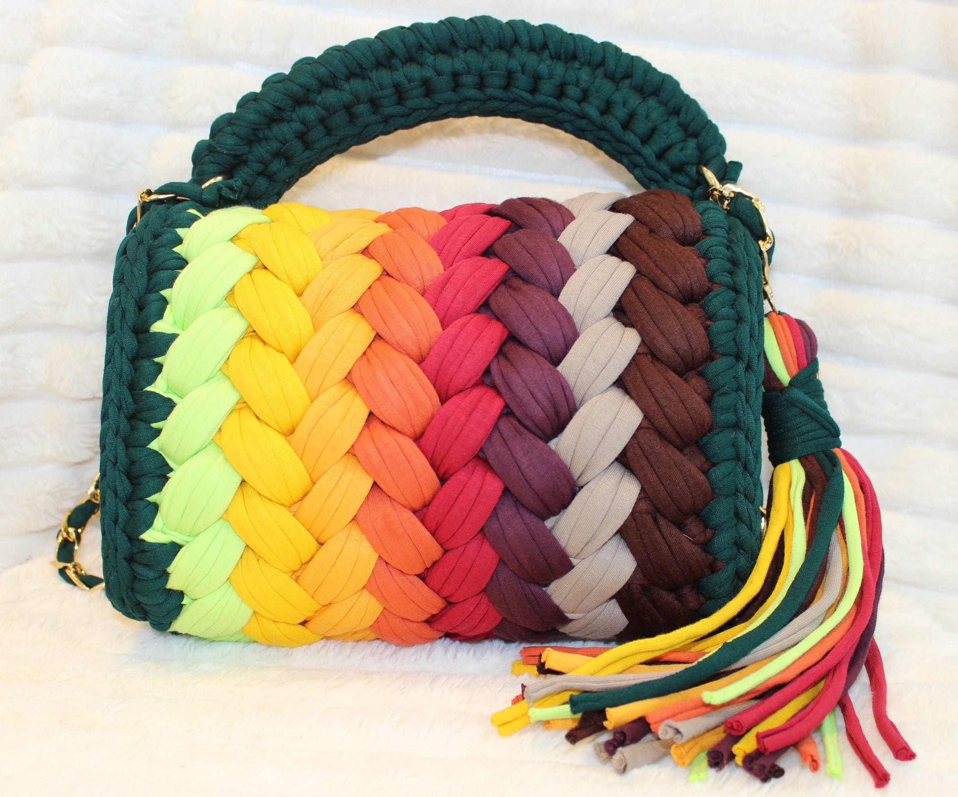 Back view of bag in natural lighting: Colorful woven Crochet handbag (Forrest Green, Red, Burgundy, Lime Green, Orange, Yellow, and Gold) with tassel sitting on a white, textured surface. 