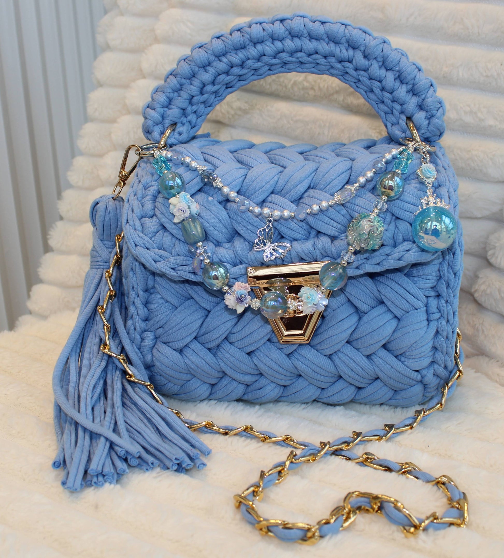 Front view of bag in natural lighting: Denim Blue woven handbag with decorative keychain and tassel on a White textured surface