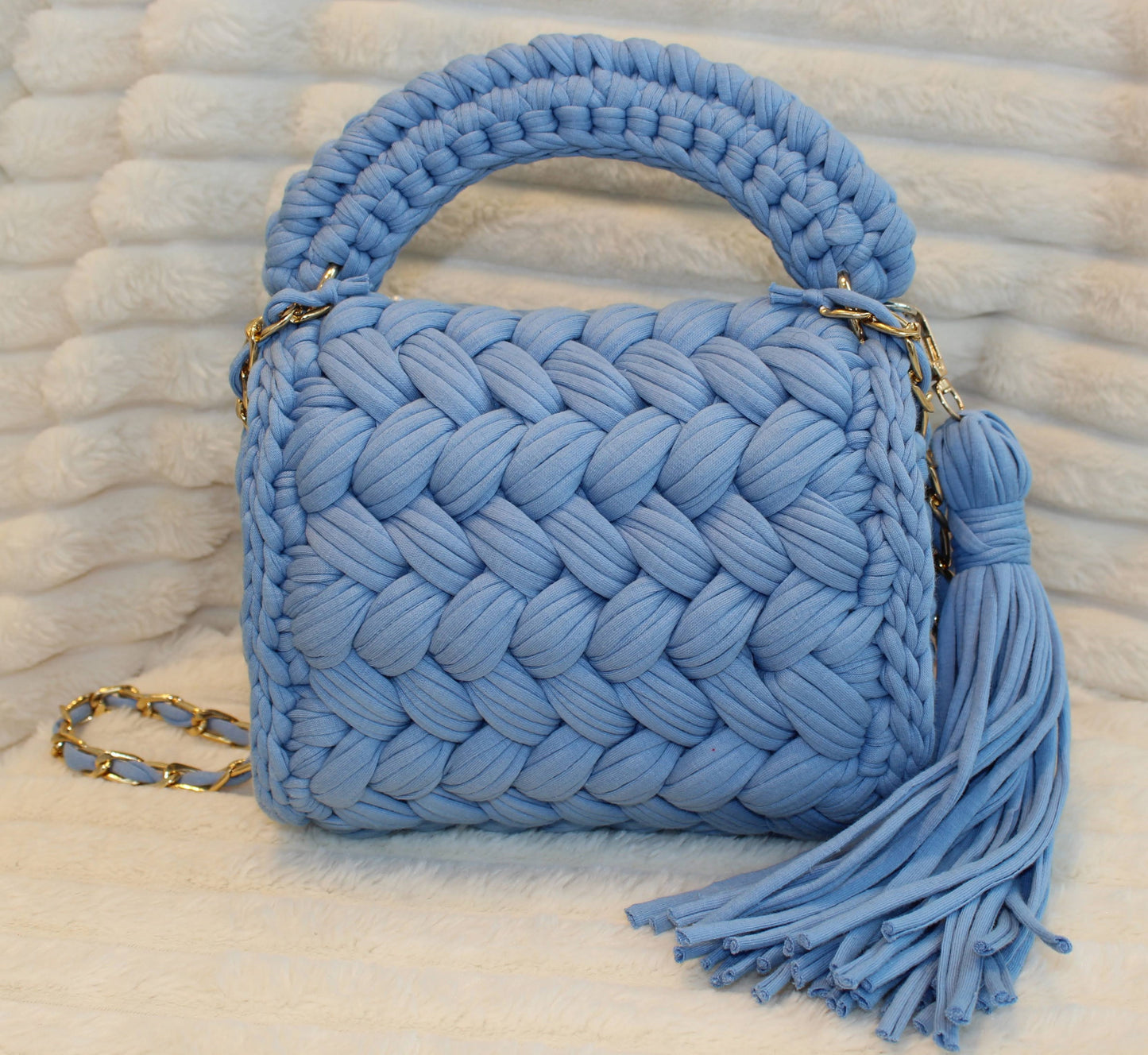 Back view of bag in natural lighting: Denim Blue woven handbag with tassel on a White textured surface