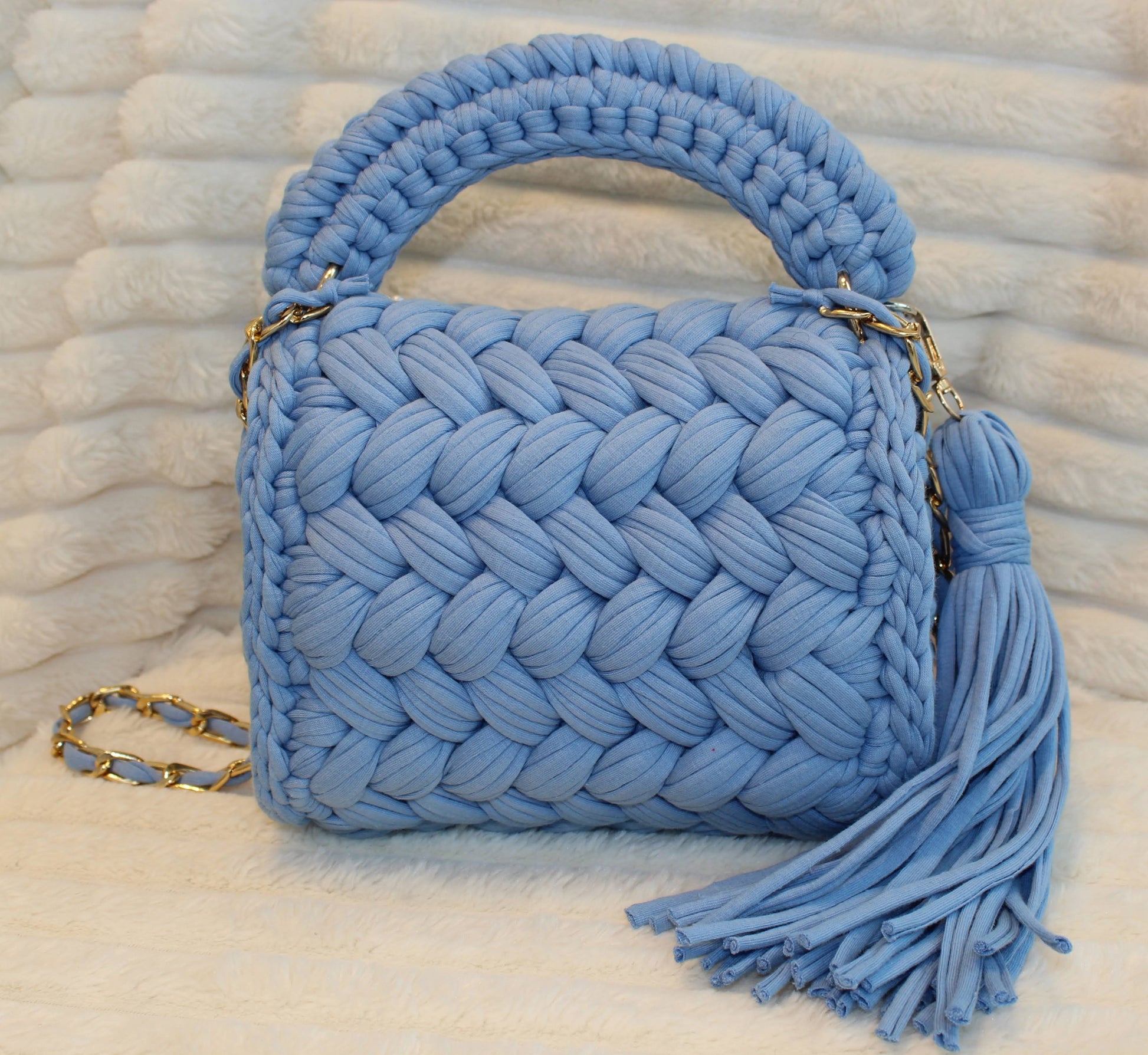 Back view of bag in natural lighting: Denim Blue woven handbag with tassel on a White textured surface