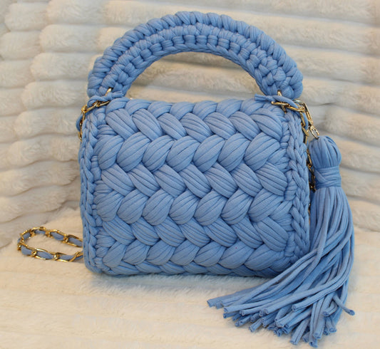 Back view of bag in natural lighting: Denim Blue woven handbag with tassel on a White textured surface