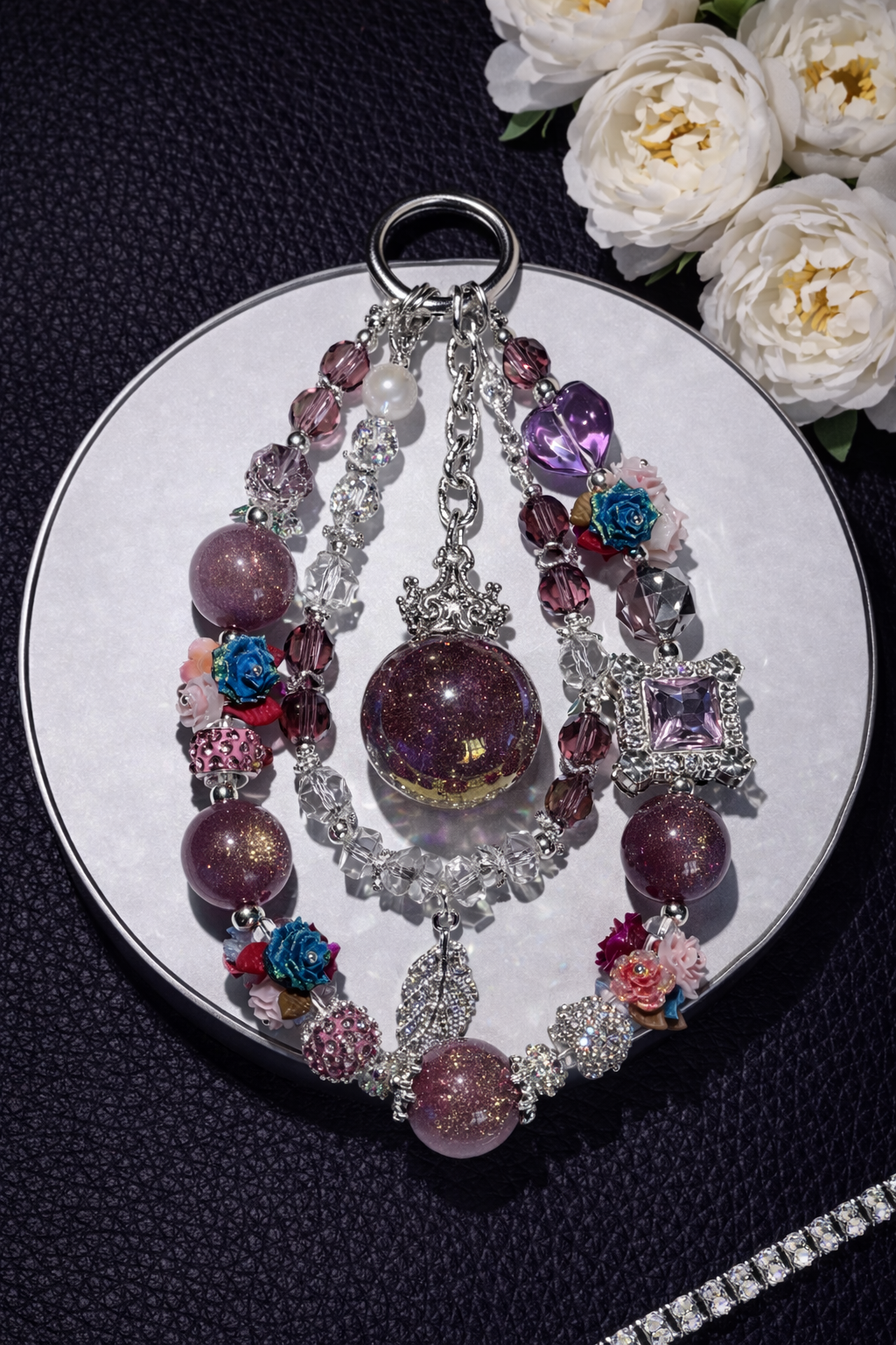 Decorative silver keychain with shimmering plum beads, colorful floral accents, and a feather pendant on a black leather surface with white flowers in the top right corner and a silver chain in the bottom right corner. 