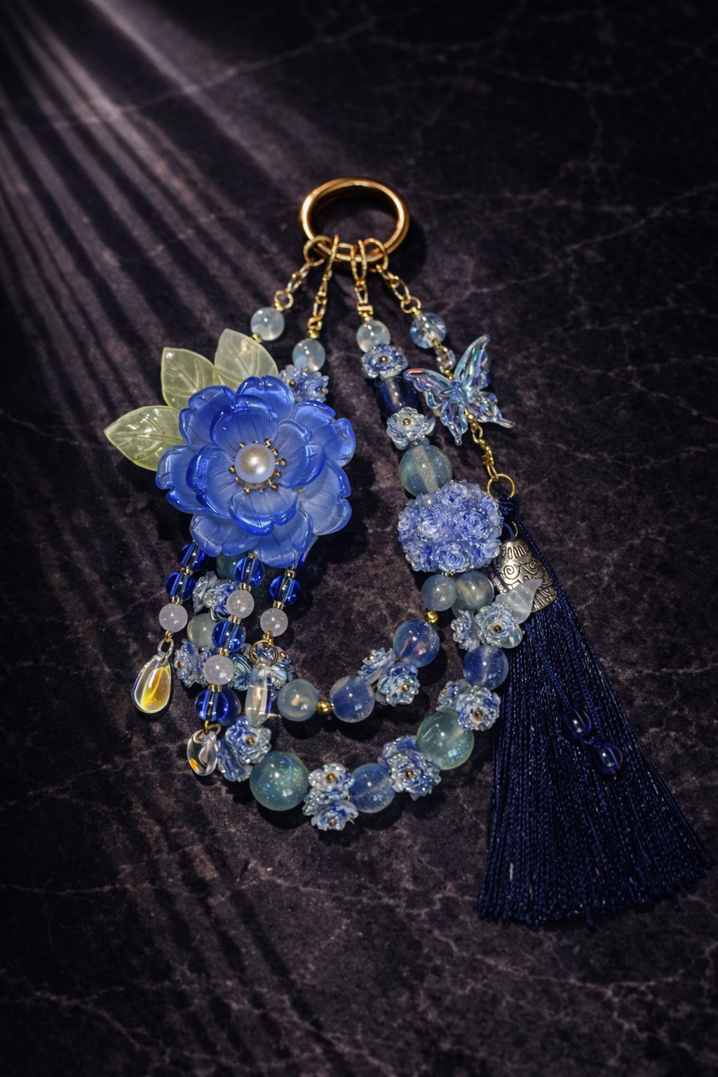 Decorative blue keychain with a large blue flower charm, light blue shimmering beads, floral accents, and a dark blue tassel on a dark marble background