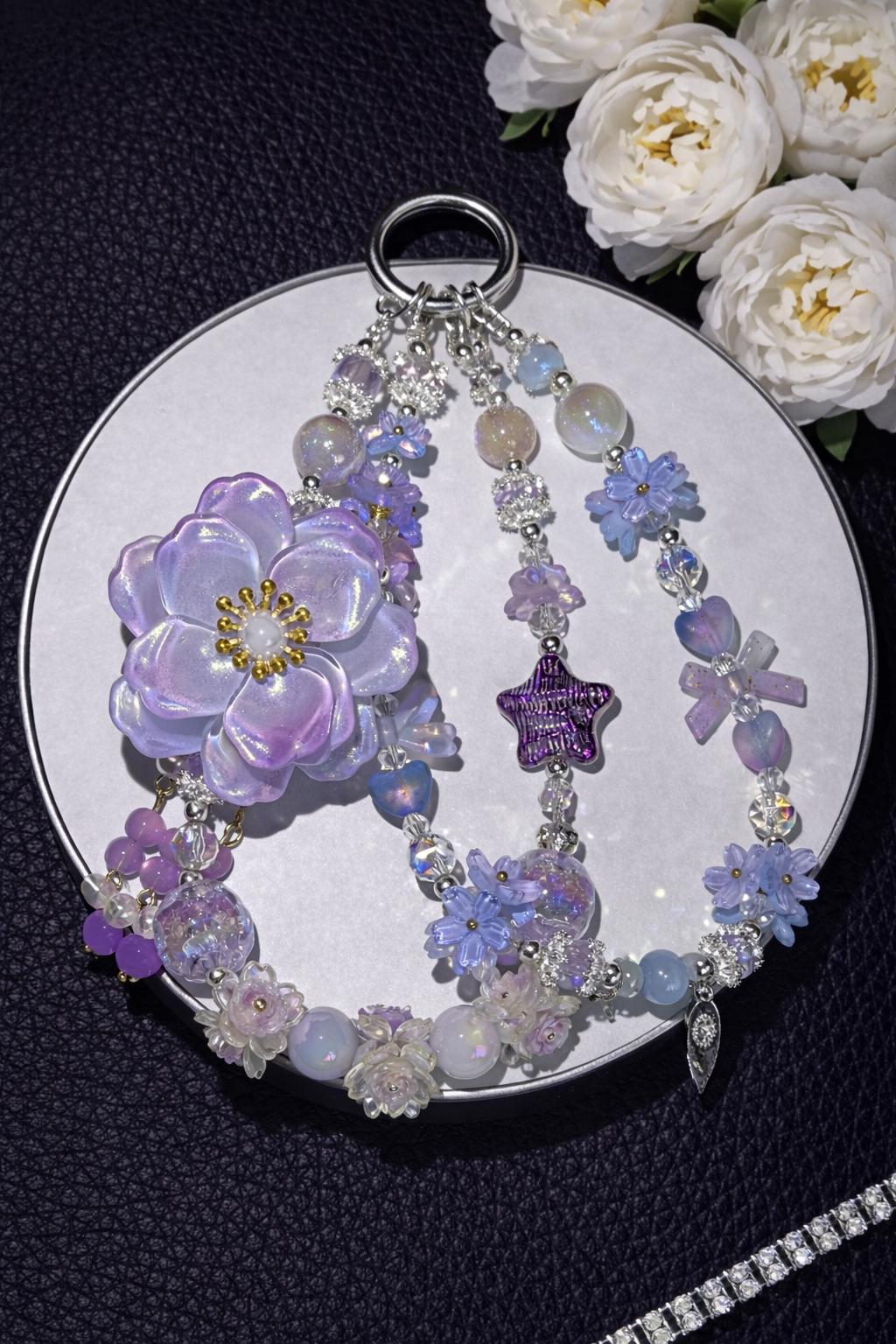 Decorative keychain with lilac and lavender flower beads, a large lilac flower charm, and shimmering accents on a black leather surface with flowers in the top right corner and a silver chain in the bottom right corner. 