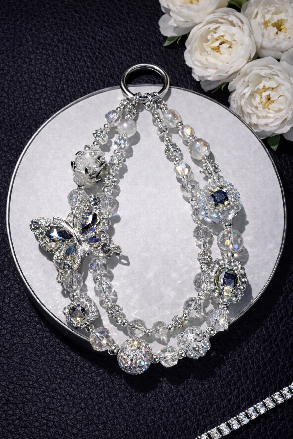 Decorative silver keychain with sparkling beads, a large butterfly charm, and shimmering accents on a black leather surface with white flowers in the top right corner and a silver chain in the bottom right corner. 