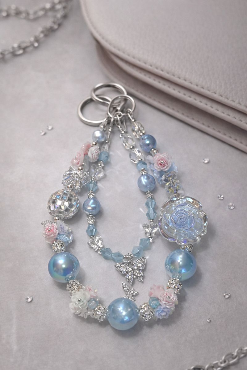 Decorative silver keychain with shimmering blue beads, disco charms, floral accents, and a silver butterfly pendant on soft, beige surface with crystal decor.